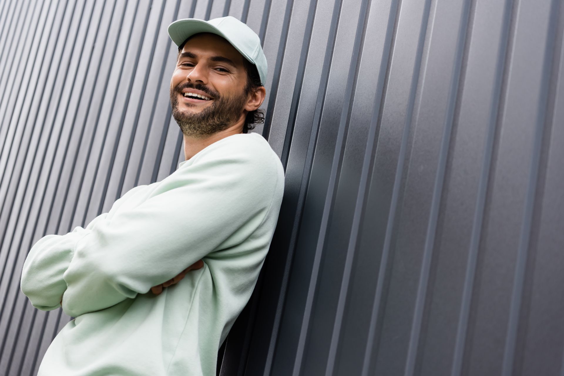 Un homme souriant, vêtu d'un sweat-shirt et d'une casquette vert clair, se tient debout, les bras croisés, contre un mur sombre en tôle ondulée.