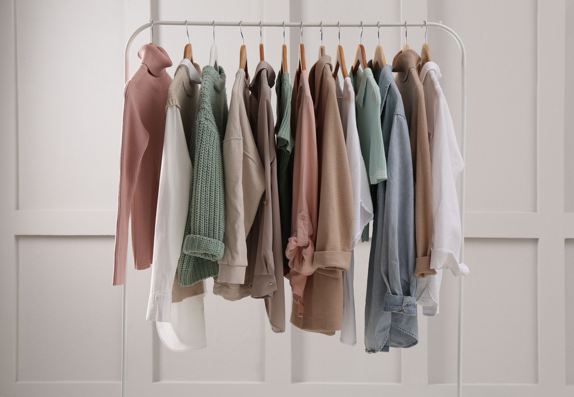 Des pulls et des vestes aux tons pastel sont suspendus à un portant blanc devant un mur clair.