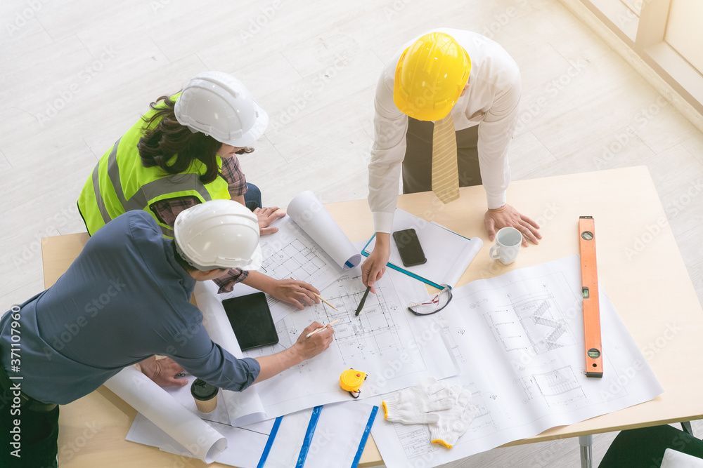 Trois ouvriers du bâtiment, casqués, examinent des plans à une table où se trouvent des outils et des plans.