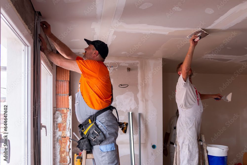 Deux ouvriers appliquent de l'enduit à plaques de plâtre sur un plafond lors de la rénovation d'une maison.