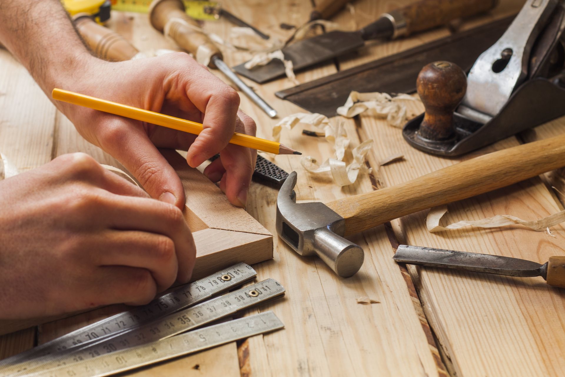 Des mains sculptent du bois avec un crayon et un ciseau sur un établi, entourées d'outils et de copeaux de bois.