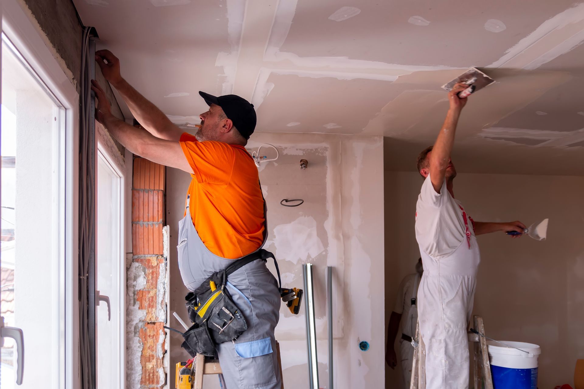 Deux ouvriers enduisent et poncent le plafond d'une pièce lors d'une rénovation intérieure.