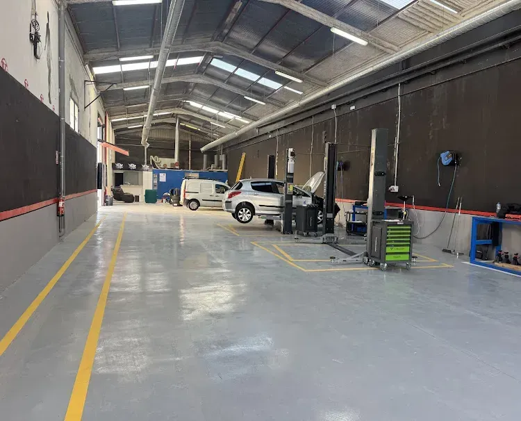 Interior de un taller mecánico con un vehículo en un elevador. Suelo gris, techos altos y herramientas visibles.
