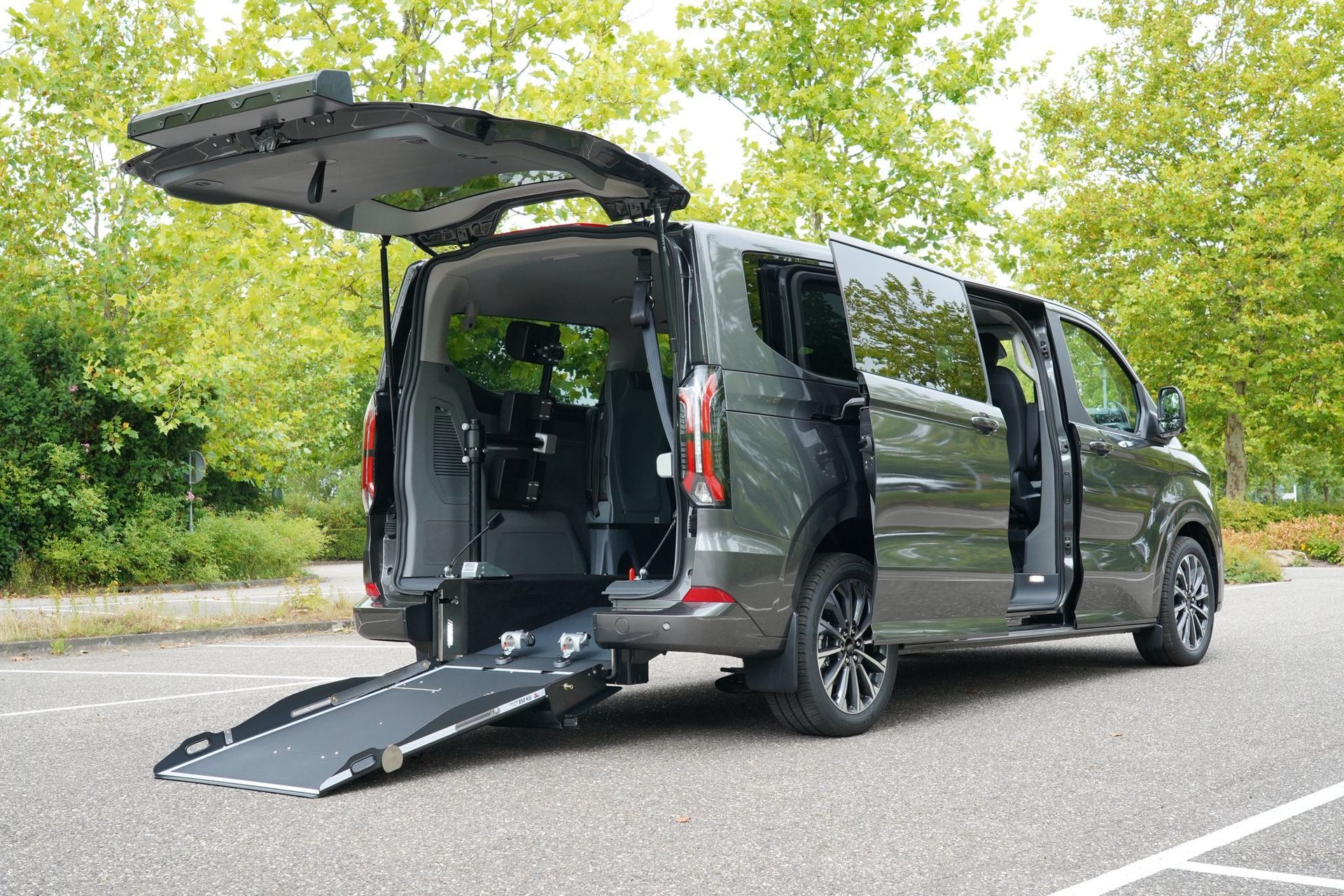 Fourgonnette accessible gris foncé avec rampe déployée, porte arrière ouverte et porte latérale coulissante, stationnée sur un parking.