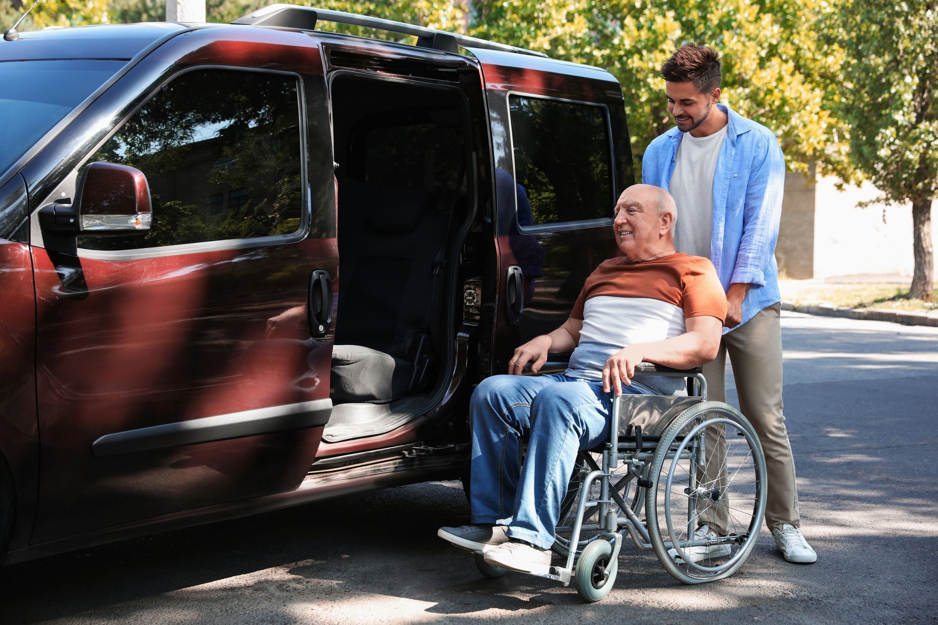 Un homme en fauteuil roulant, assis à côté d'une camionnette dont une porte latérale est ouverte, est aidé par une autre personne dans la rue.