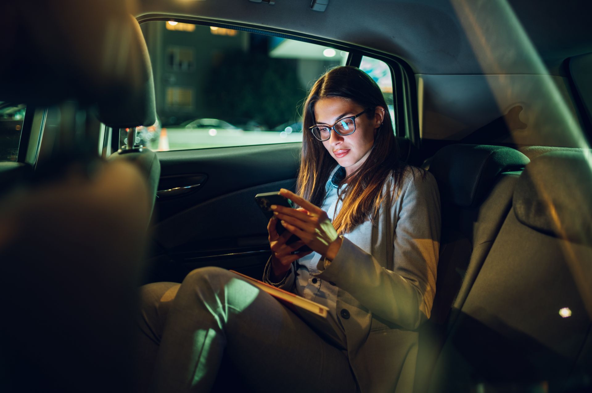 Une femme portant des lunettes utilise un smartphone sur la banquette arrière d'une voiture, la nuit.