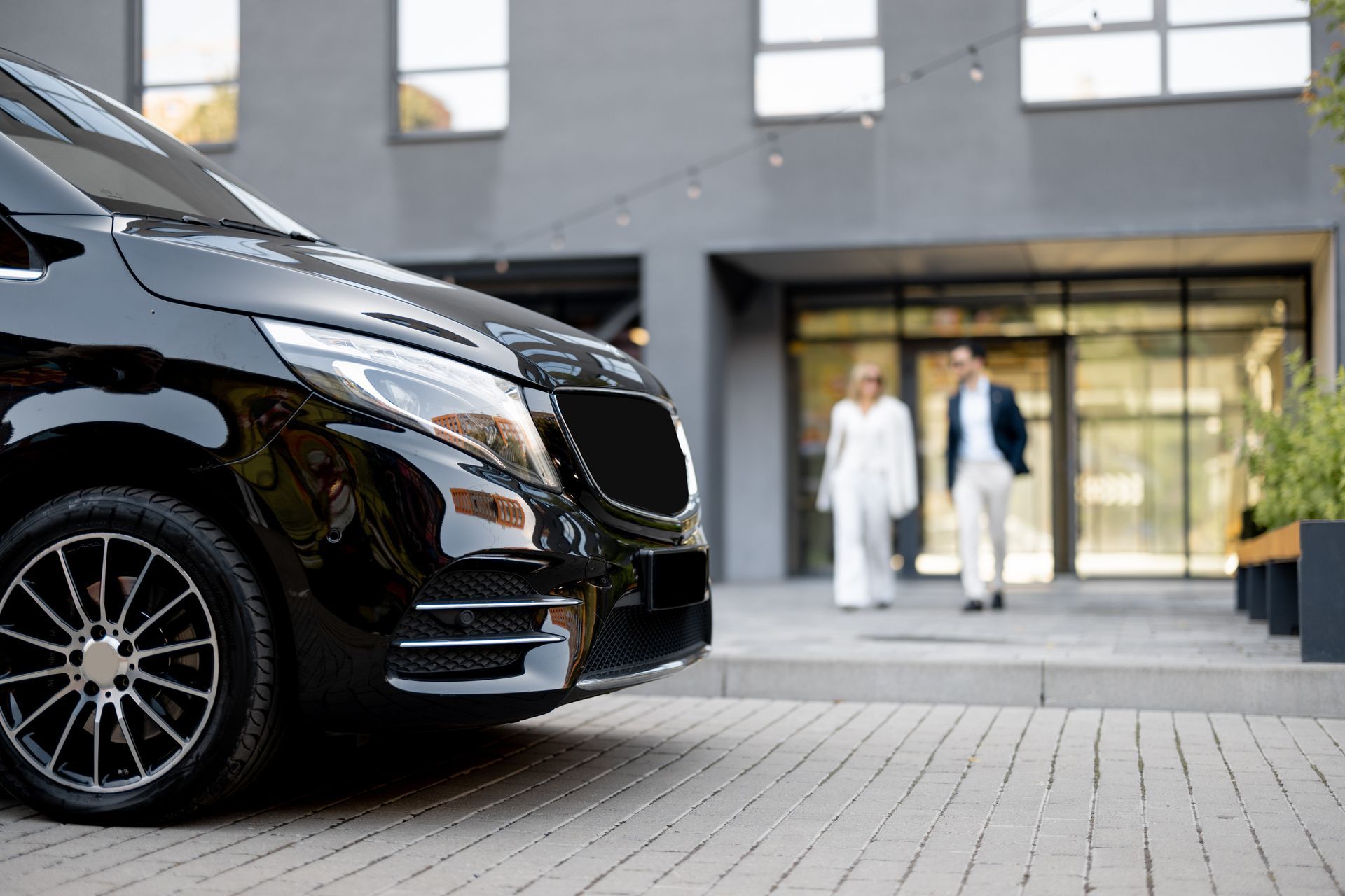 Une voiture de luxe noire est garée devant un immeuble moderne, deux personnes se tenant à l'entrée.