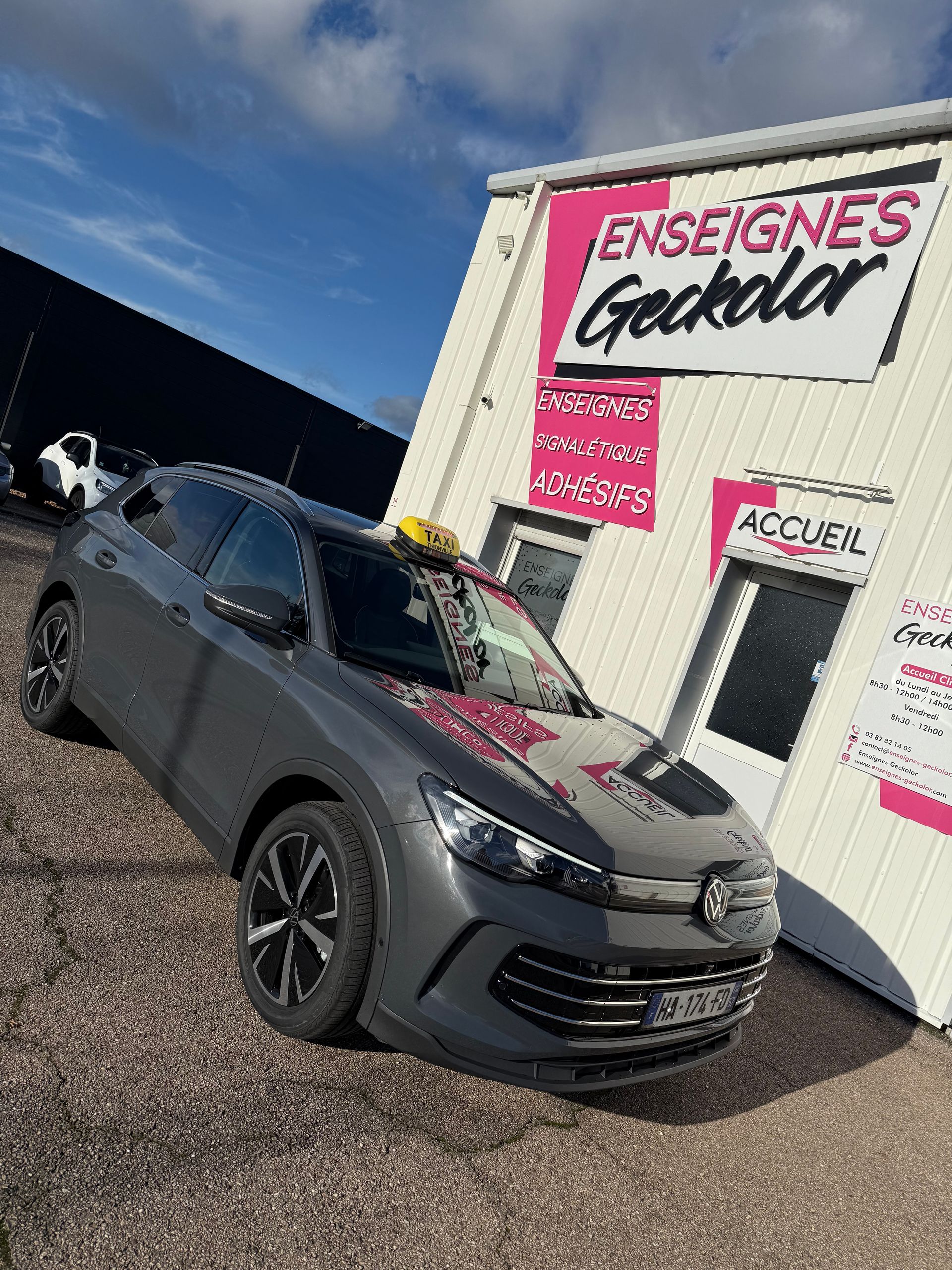 Voiture grise garée devant un bâtiment à l'enseigne rose, ciel bleu.