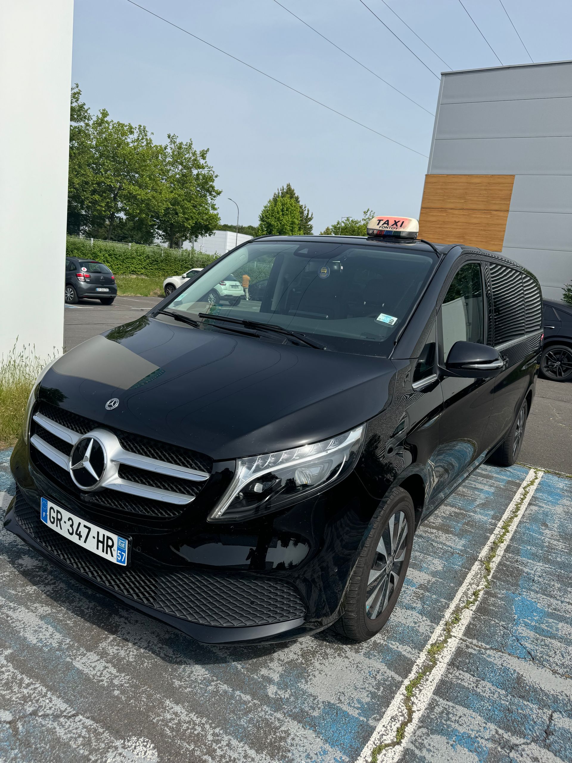 Un taxi Mercedes-Benz Classe V noir est stationné sur un parking sous un ciel partiellement nuageux.