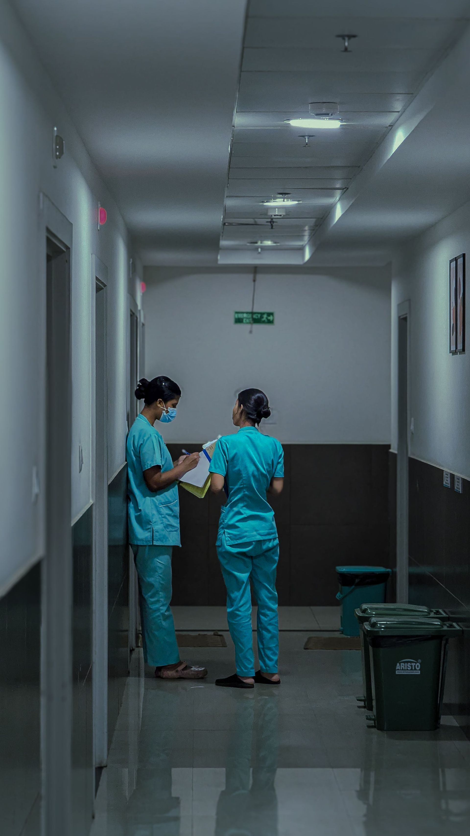 Deux membres du personnel médical en blouse turquoise discutent dans un couloir d'hôpital sous des néons.