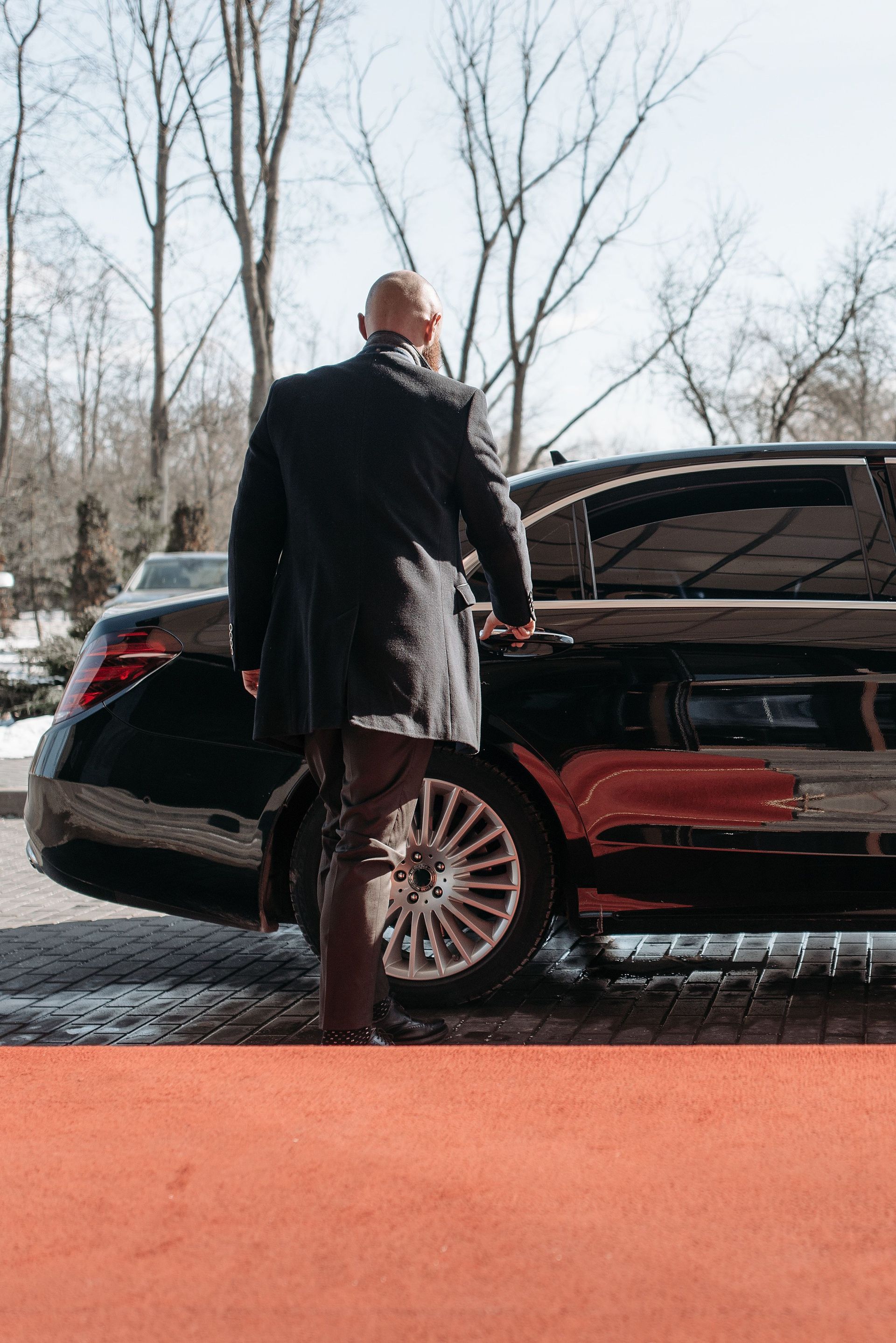 Un homme en manteau sombre marche vers une berline noire sur un tapis rouge à l'extérieur en hiver