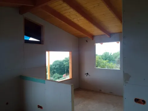 Habitación vacía en construcción, con vigas de madera en el techo y ventanas abiertas con vistas a los árboles