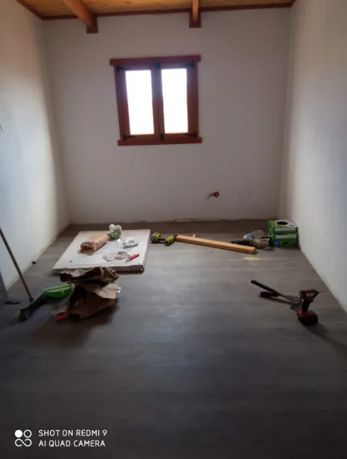 Habitación vacía en proceso de reforma, con herramientas y materiales en el suelo, paredes blancas, una ventana de madera y suelo gris.