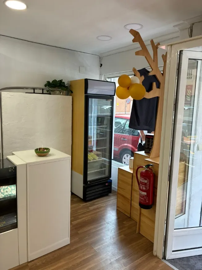 Interior de una pequeña tienda con vitrinas, una nevera de cristal para bebidas y globos amarillos colgando cerca de la entrada.