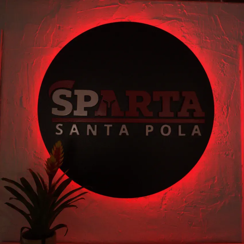 Letrero circular de Sparta Santa Pola iluminado en rojo sobre una pared con la silueta de una planta.