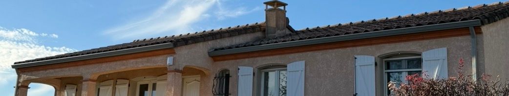 Un bâtiment au toit de tuiles, aux volets blancs et à la cheminée, se détachant sur un ciel bleu.
