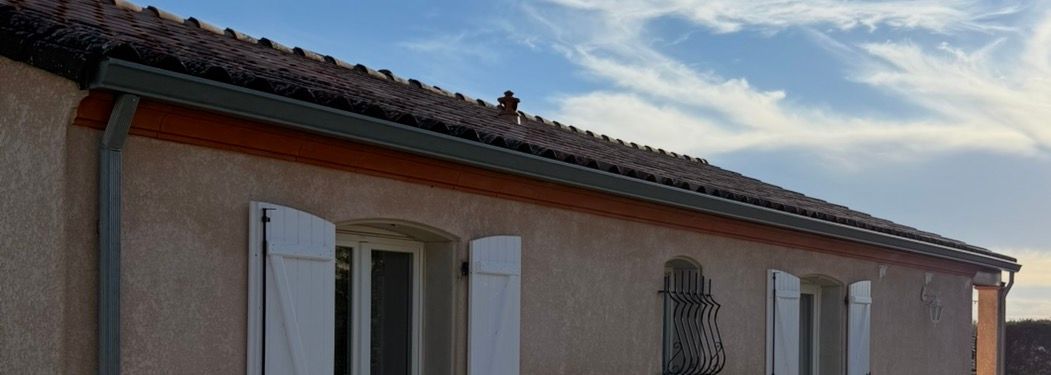 Extérieur d'un bâtiment beige avec un toit de tuiles rouges, des volets blancs et un ciel bleu nuageux.