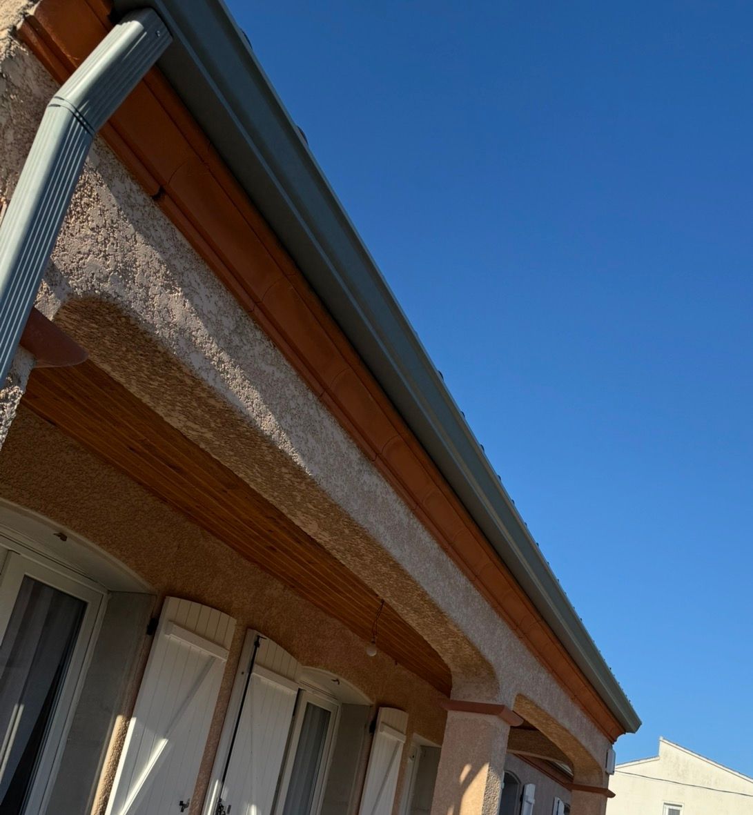 Extérieur d'une maison aux gouttières grises, aux boiseries brunes et au crépi beige, sur fond de ciel bleu.