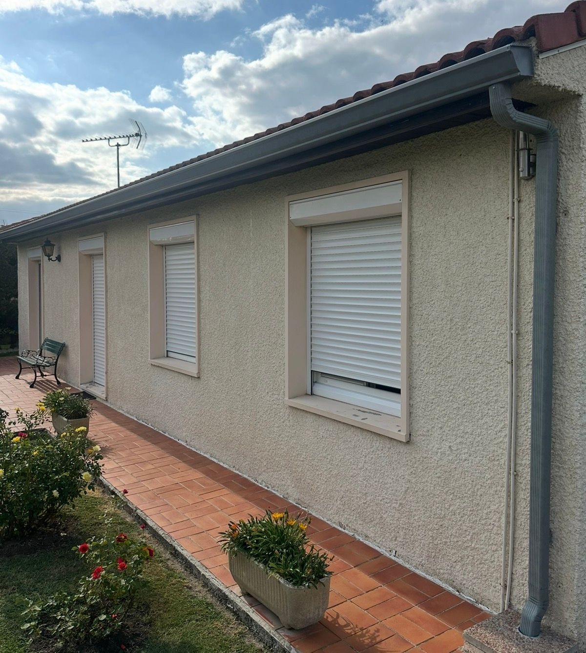 Maison beige aux gouttières grises et trois fenêtres à volets blancs. Allée en briques rouges et jardin verdoyant.