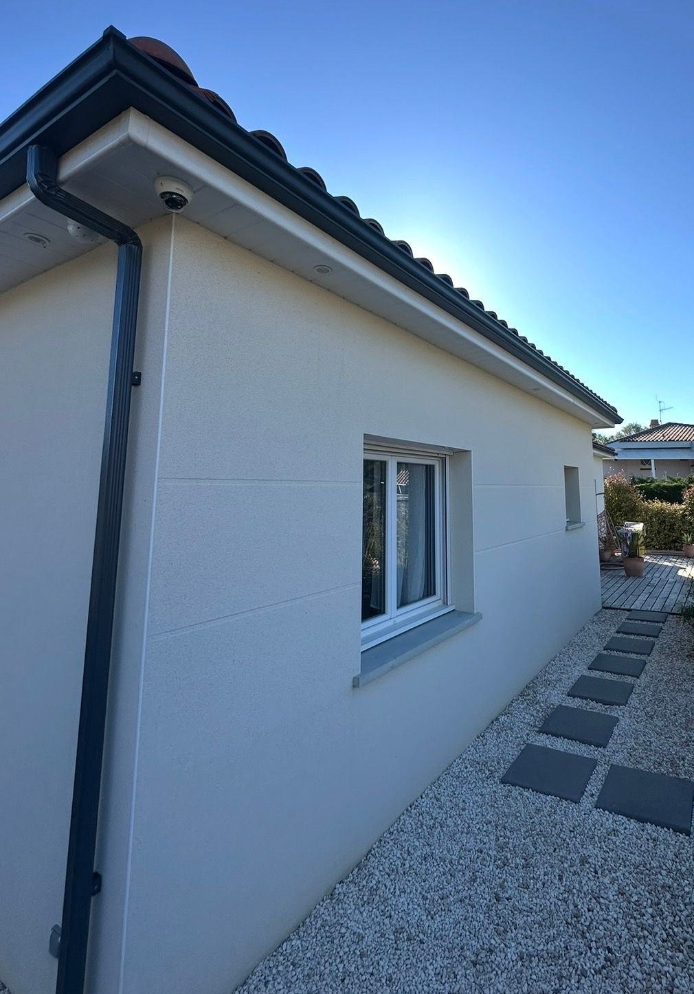 Extérieur d'une maison aux murs en stuc clair, au toit et aux boiseries foncées, et dotée d'une petite fenêtre.
