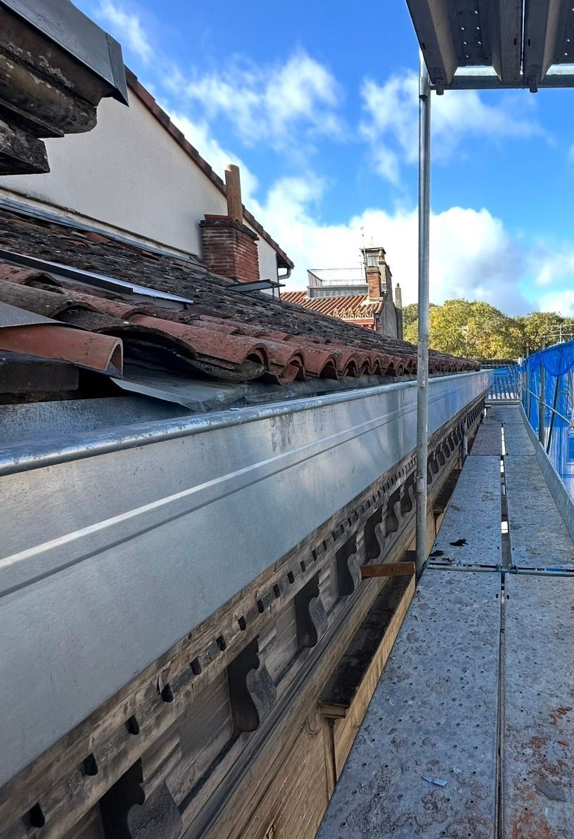 Toit avec gouttière métallique, supports en bois, échafaudage et ciel bleu.
