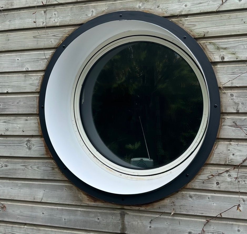 Fenêtre ronde avec encadrement noir et blanc dans un mur en bois gris.