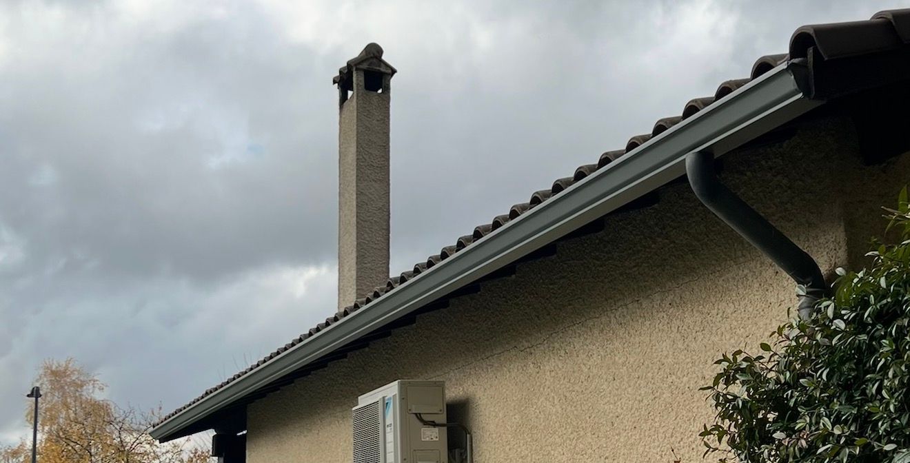 Toit gris avec gouttière, cheminée et mur en stuc sur fond de ciel nuageux.
