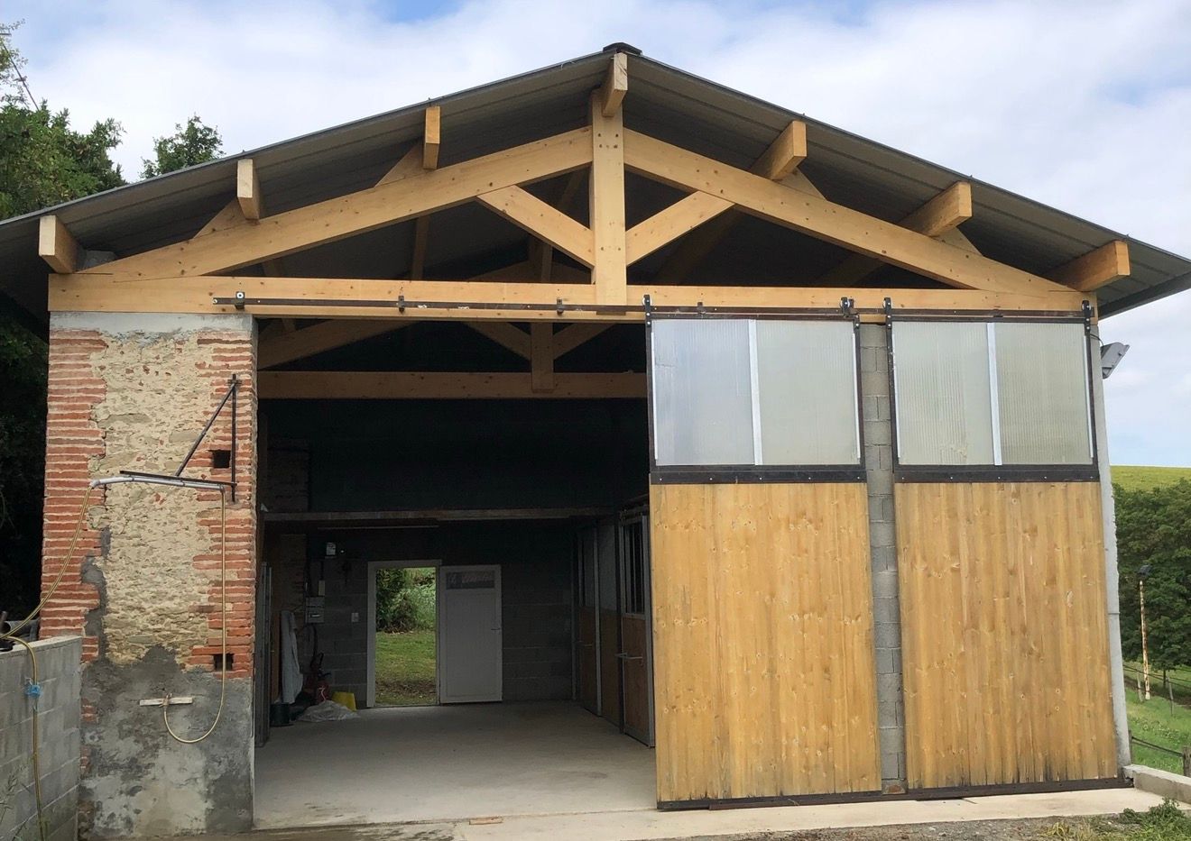 Abri de jardin en bois avec portes coulissantes, fondations en briques et béton, porte ouverte, et arrière-plan composé d'herbe et d'arbres.