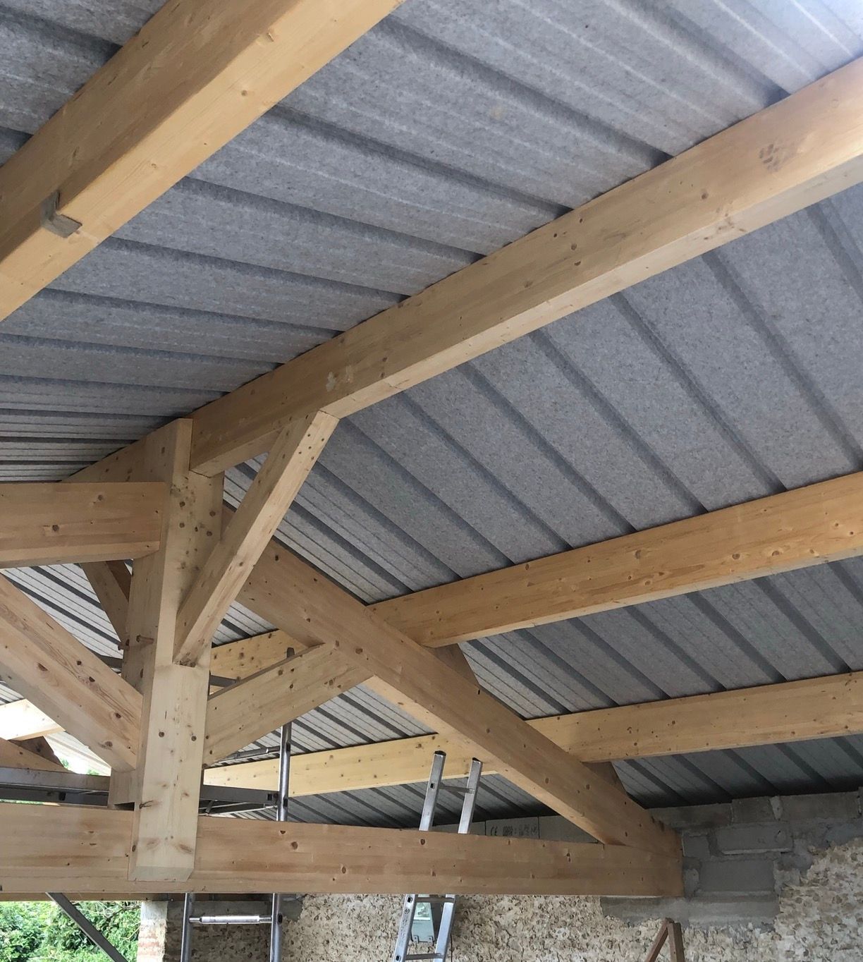 Poutres de charpente en bois soutenant une toiture en tôle ondulée grise. Vue intérieure.