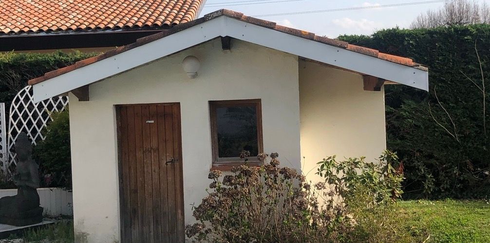 Un petit bâtiment couleur crème avec une porte et une fenêtre marron ; toit de tuiles rouges visible.