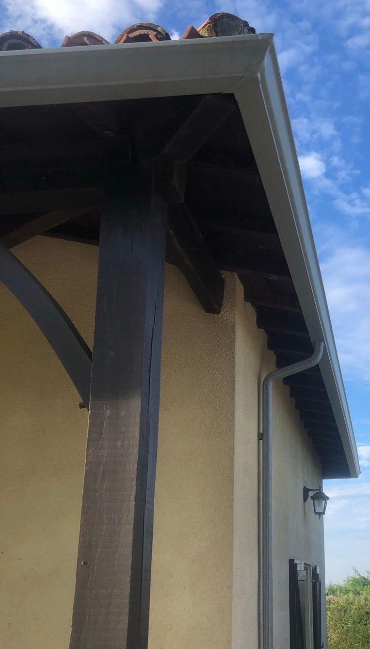 Coin d'un bâtiment avec gouttière, poutres en bois sombre et mur en stuc sur fond de ciel bleu.