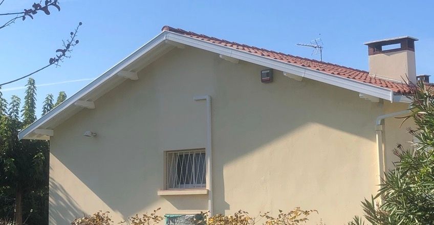 Maison beige au toit de tuiles rouges, aux boiseries blanches et à la cheminée se détachant sur un ciel bleu.