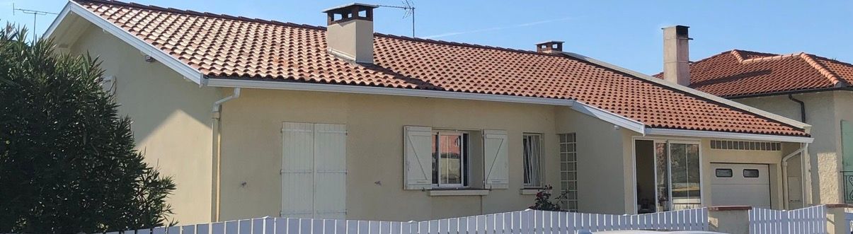 Une maison beige au toit de tuiles rouges, aux volets blancs et à la clôture blanche, sous un ciel bleu.
