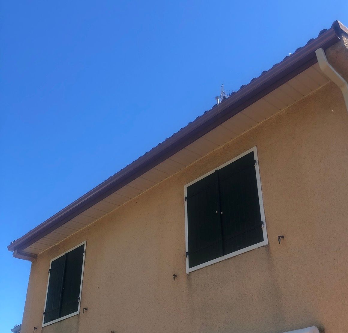Maison beige au toit et aux gouttières marron, deux fenêtres à volets noirs, sur un ciel bleu éclatant.