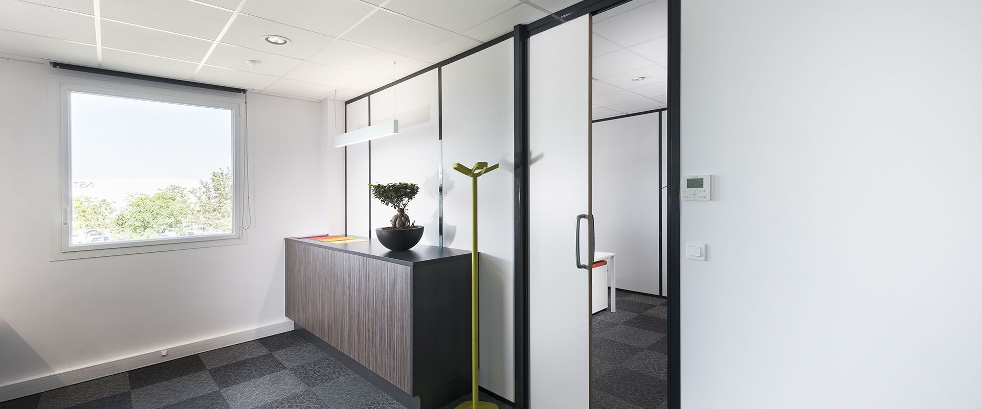 Un couloir de bureau moderne avec des murs blancs, un sol sombre, une porte vitrée et une armoire avec une plante.