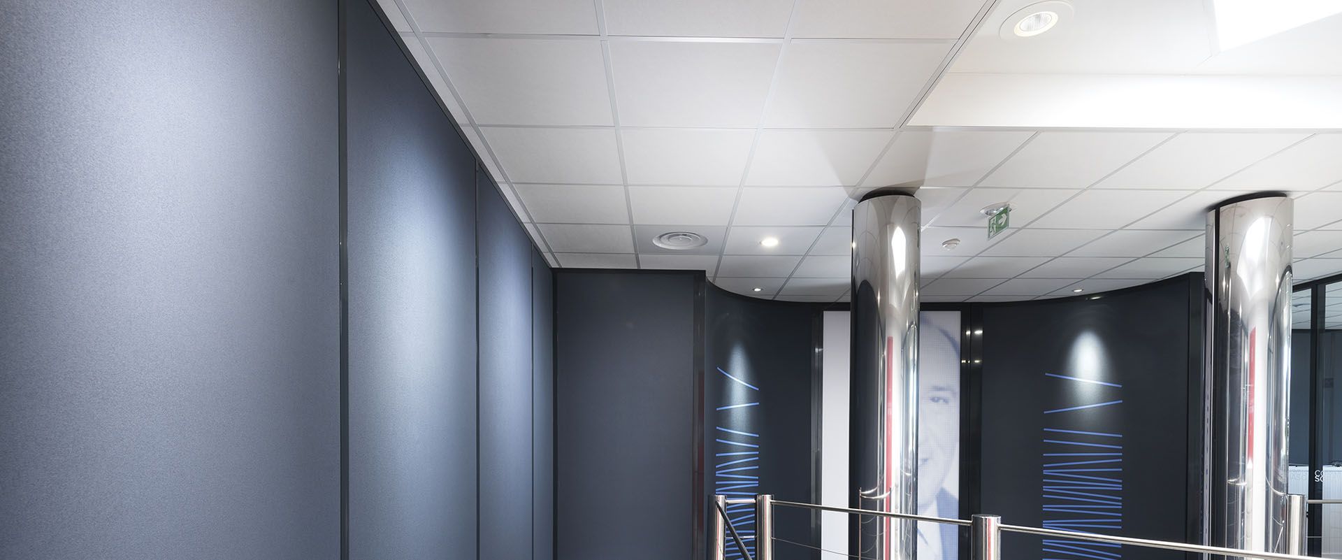 Un couloir moderne avec un mur bleu foncé, des piliers argentés et des carreaux de plafond blancs.