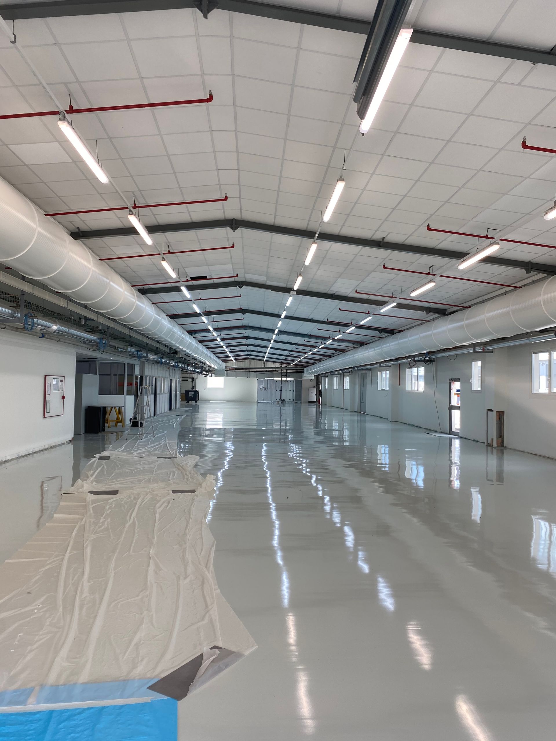 Intérieur d'un long entrepôt vide, aux murs blancs et au sol gris brillant. On aperçoit des néons et des conduits d'aération.