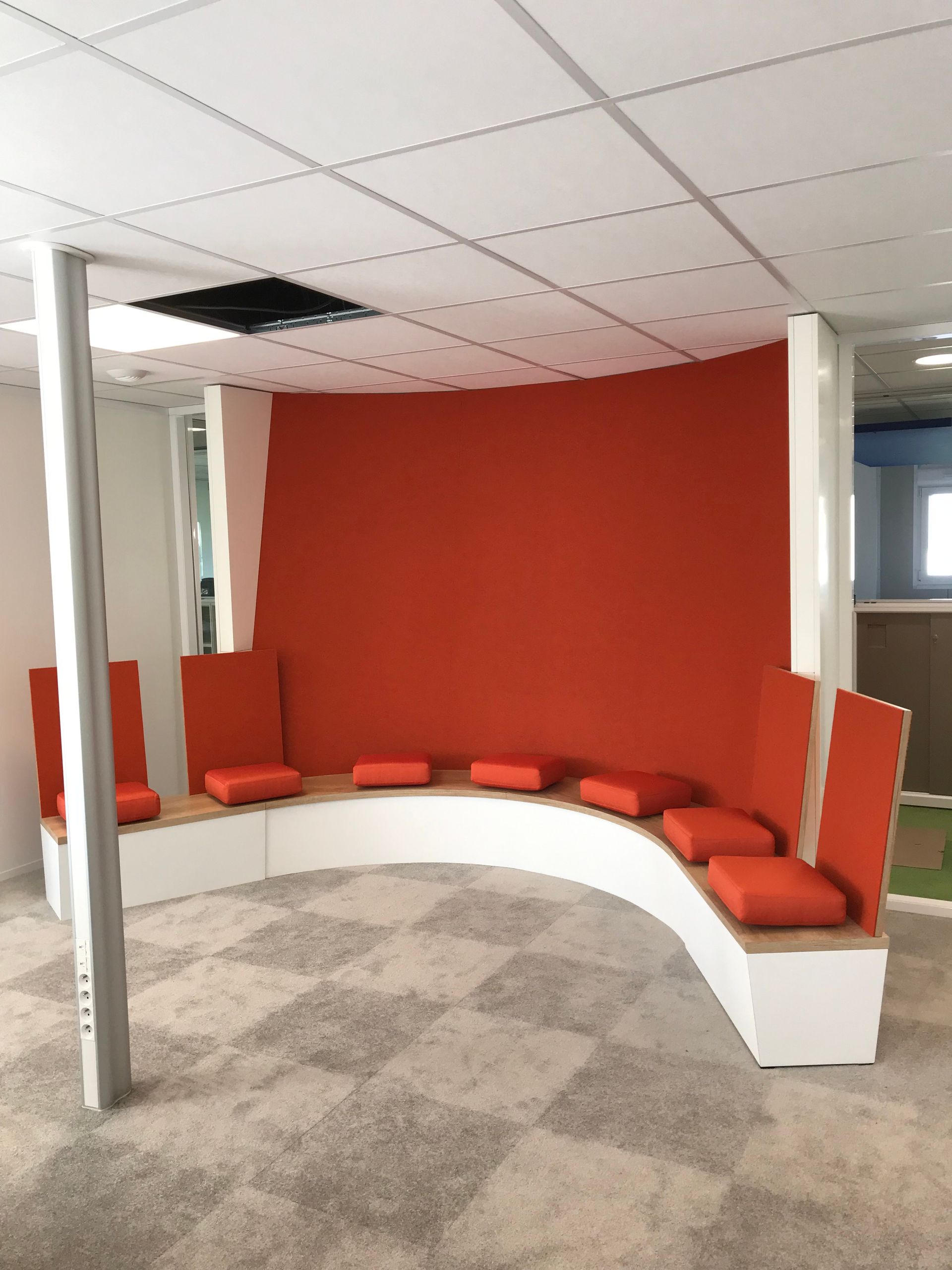 Banquette incurvée avec coussins orange et mur rouge dans une pièce à moquette à carreaux.