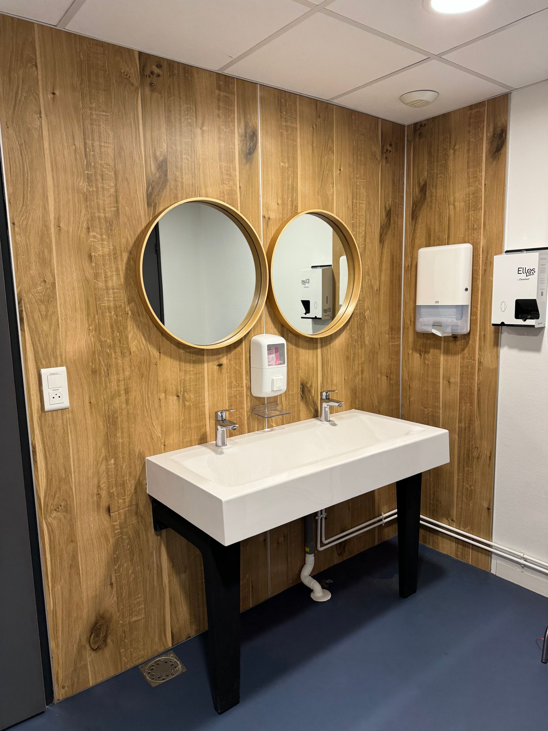 Salle de bains avec un mur lambrissé, deux miroirs ronds, un lavabo rectangulaire à deux robinets et un sol bleu.
