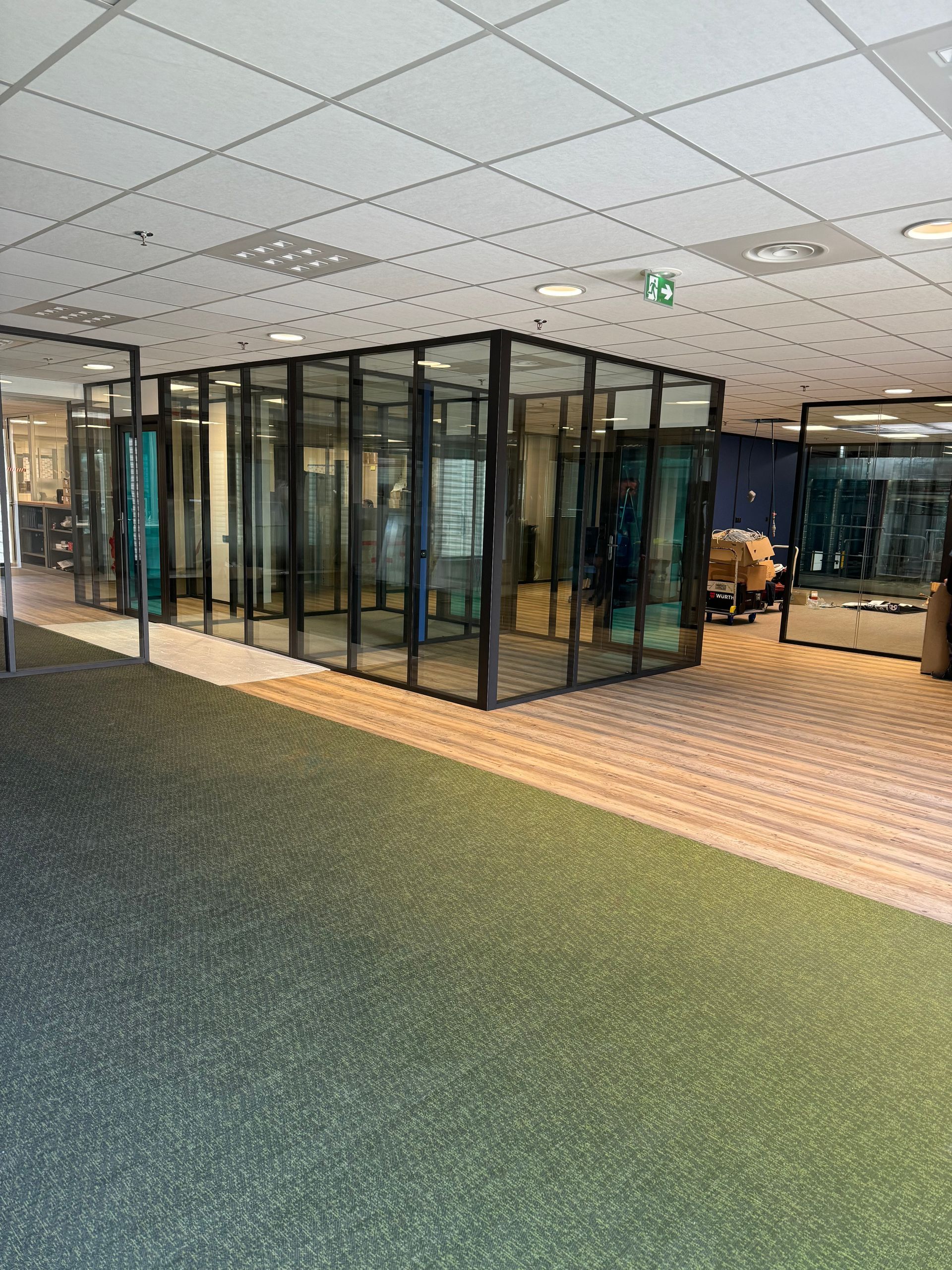 Une série de cloisons vitrées à cadres noirs dans un espace de bureaux recouvert de moquette verte.
