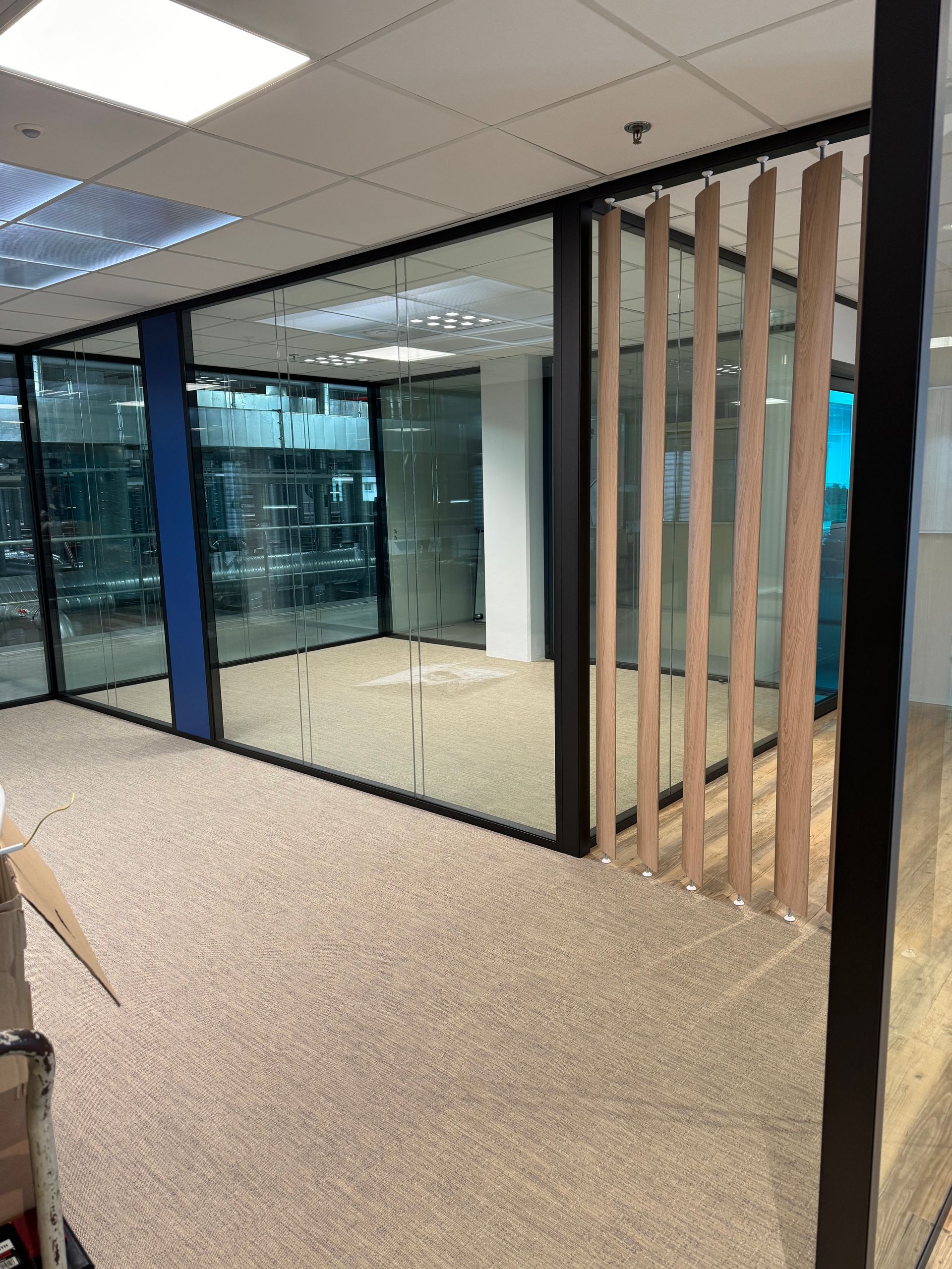 Espace de bureau vide avec murs vitrés, sol recouvert de moquette et lattes de bois verticales.