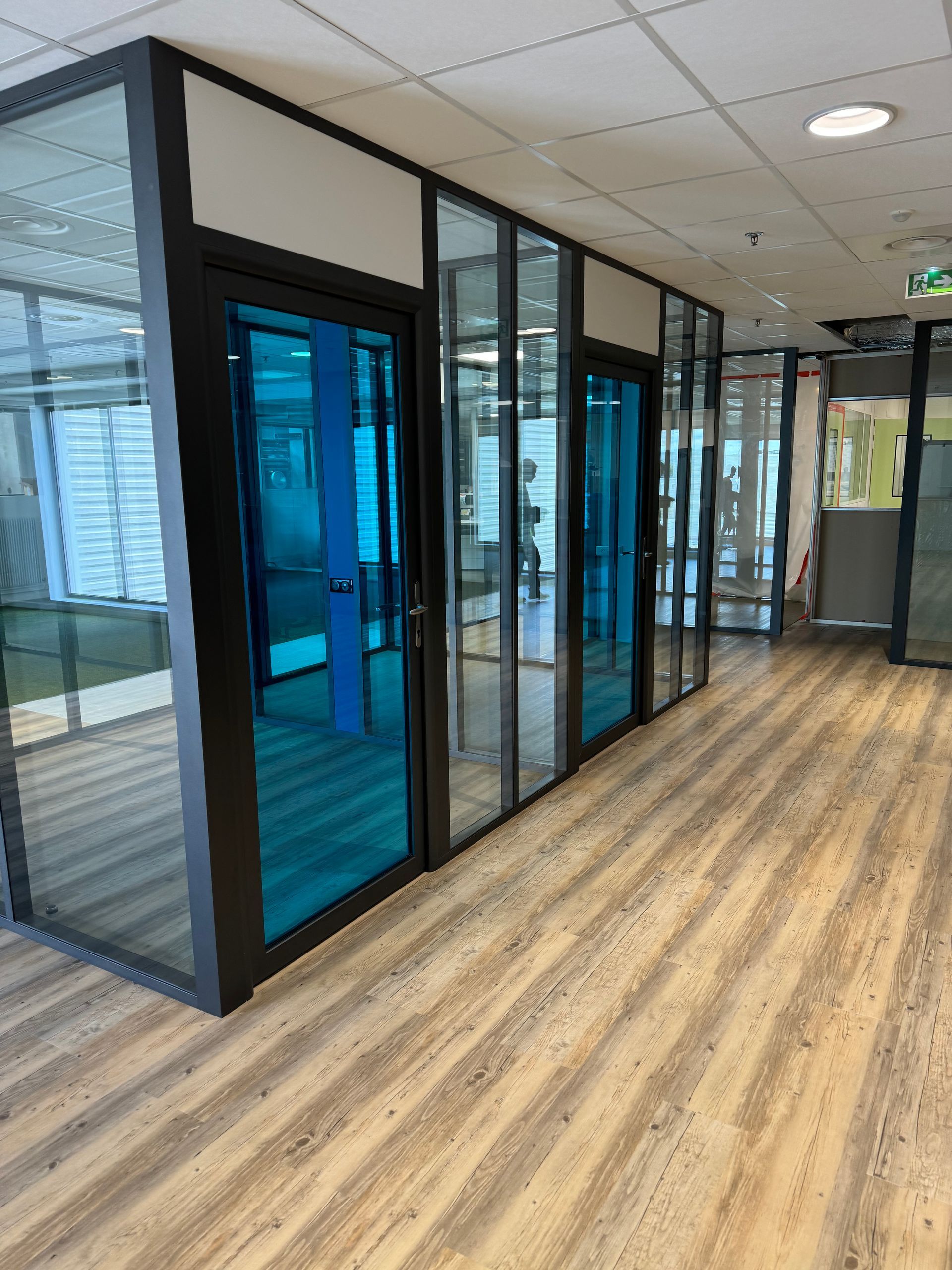Un couloir avec des bureaux aux parois vitrées. Portes teintées bleutées, cadres gris et blancs, et sol imitation bois.