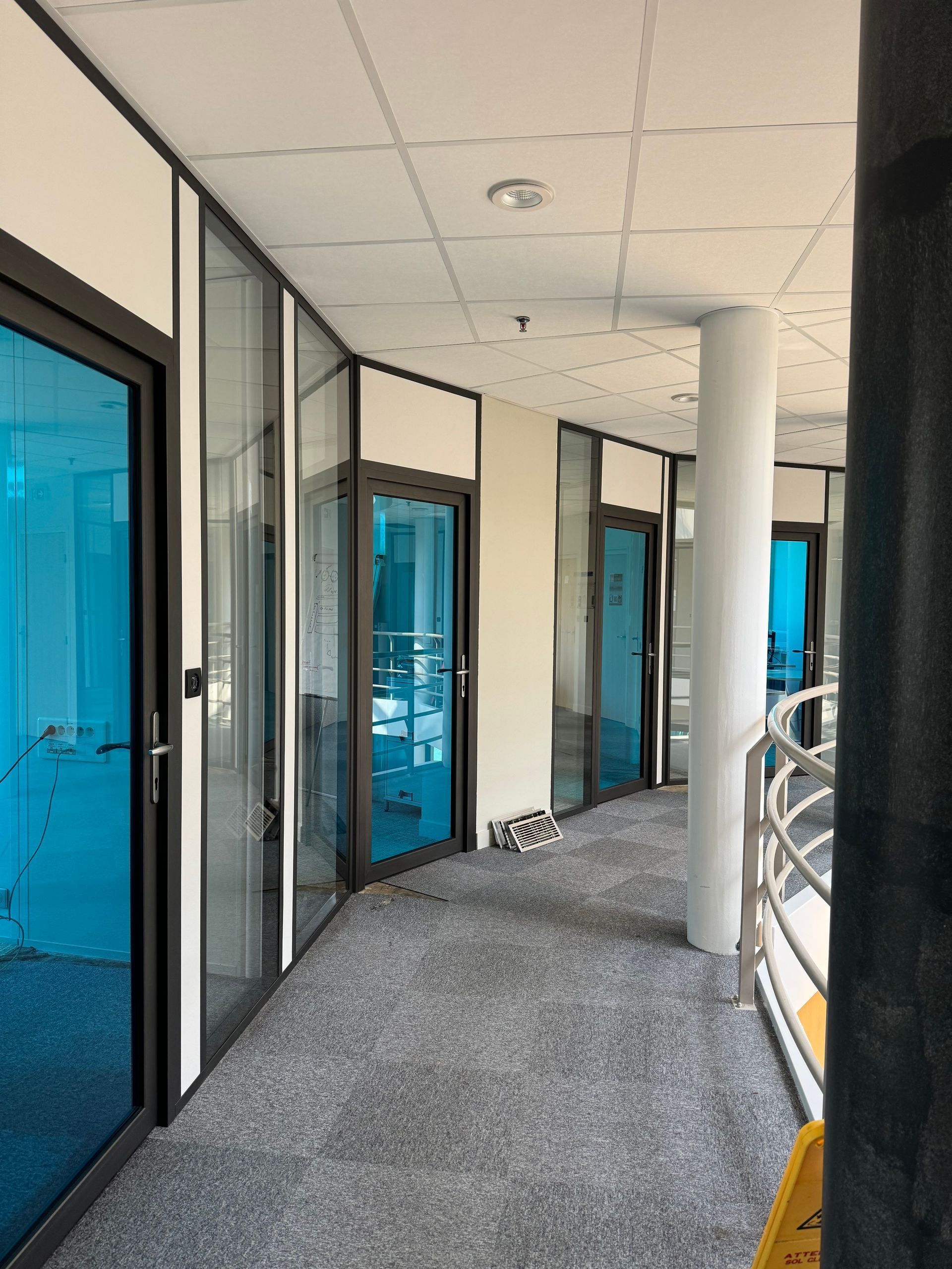 Couloir incurvé avec bureaux aux parois de verre. Portes et fenêtres teintées bleutées, moquette grise, plafond blanc.