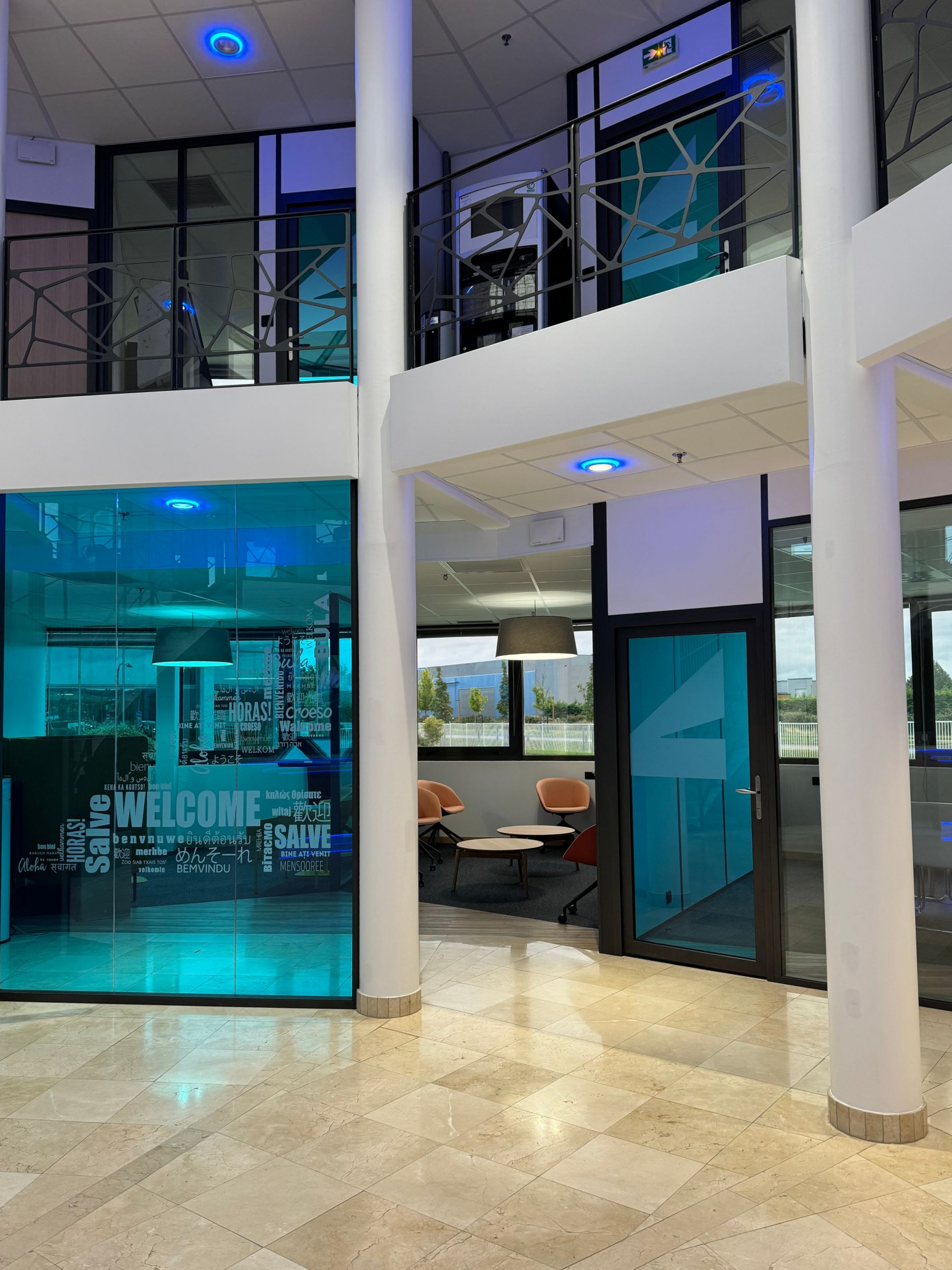 Intérieur de bureau moderne avec des murs en verre teinté bleu, une disposition sur deux étages, des colonnes blanches et un éclairage lumineux.