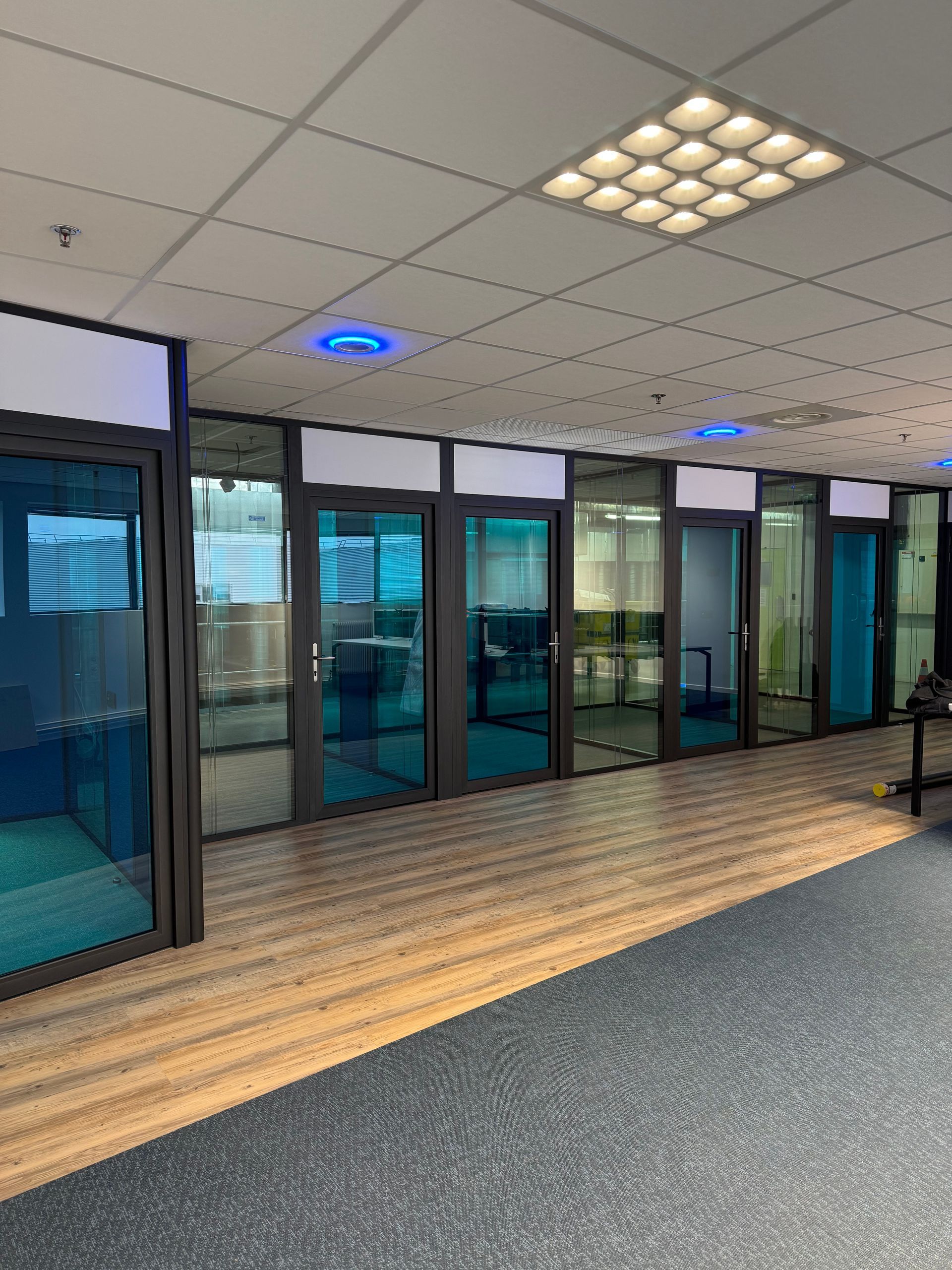 Couloir de bureau avec pièces vitrées, sol imitation bois et touches de bleu. Éclairage lumineux au plafond.