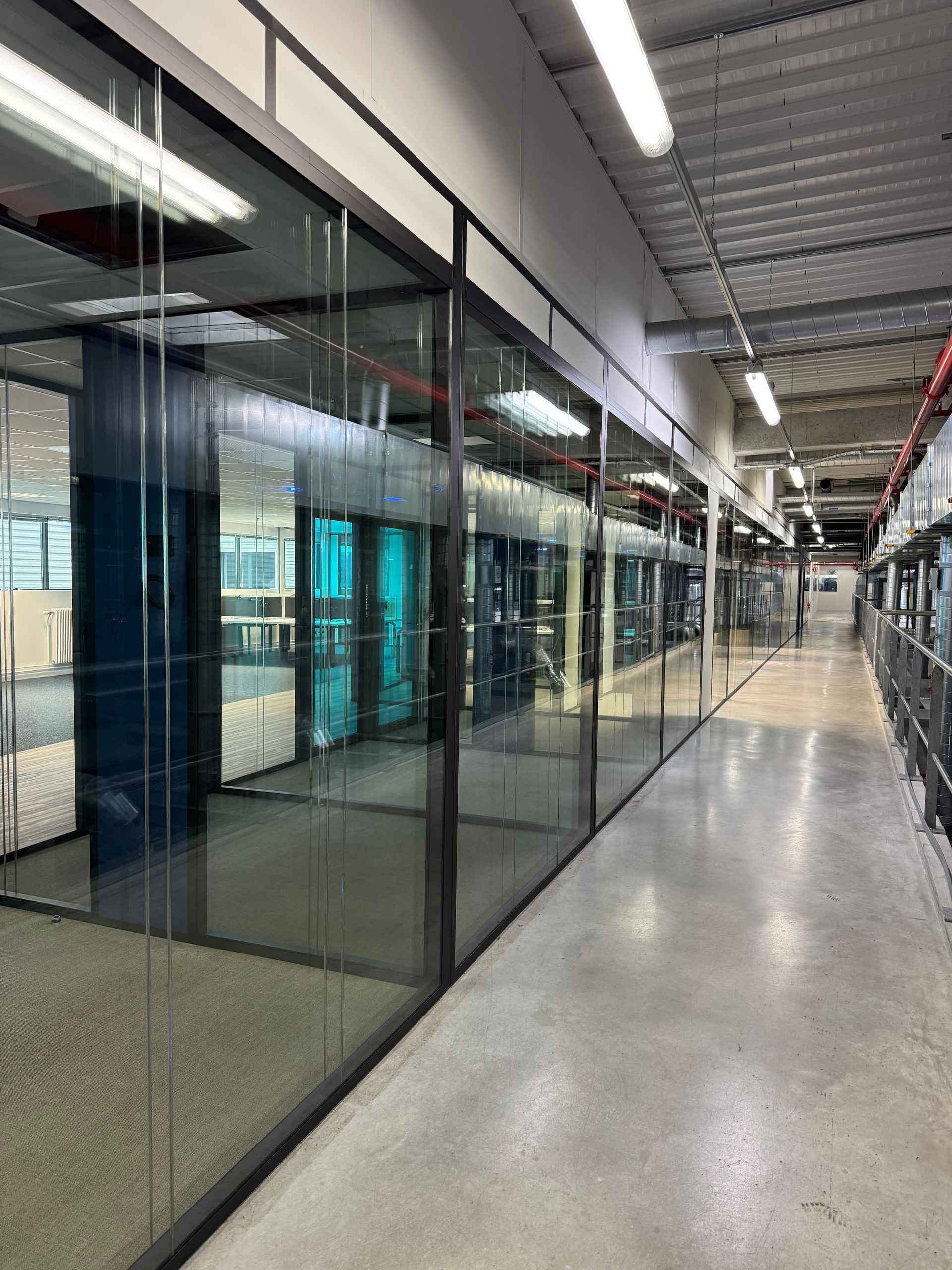 Couloir desservant des bureaux aux parois vitrées. Sol gris, plafond métallique avec éclairage intégré. Les bureaux intérieurs sont décorés de bleu et de blanc.