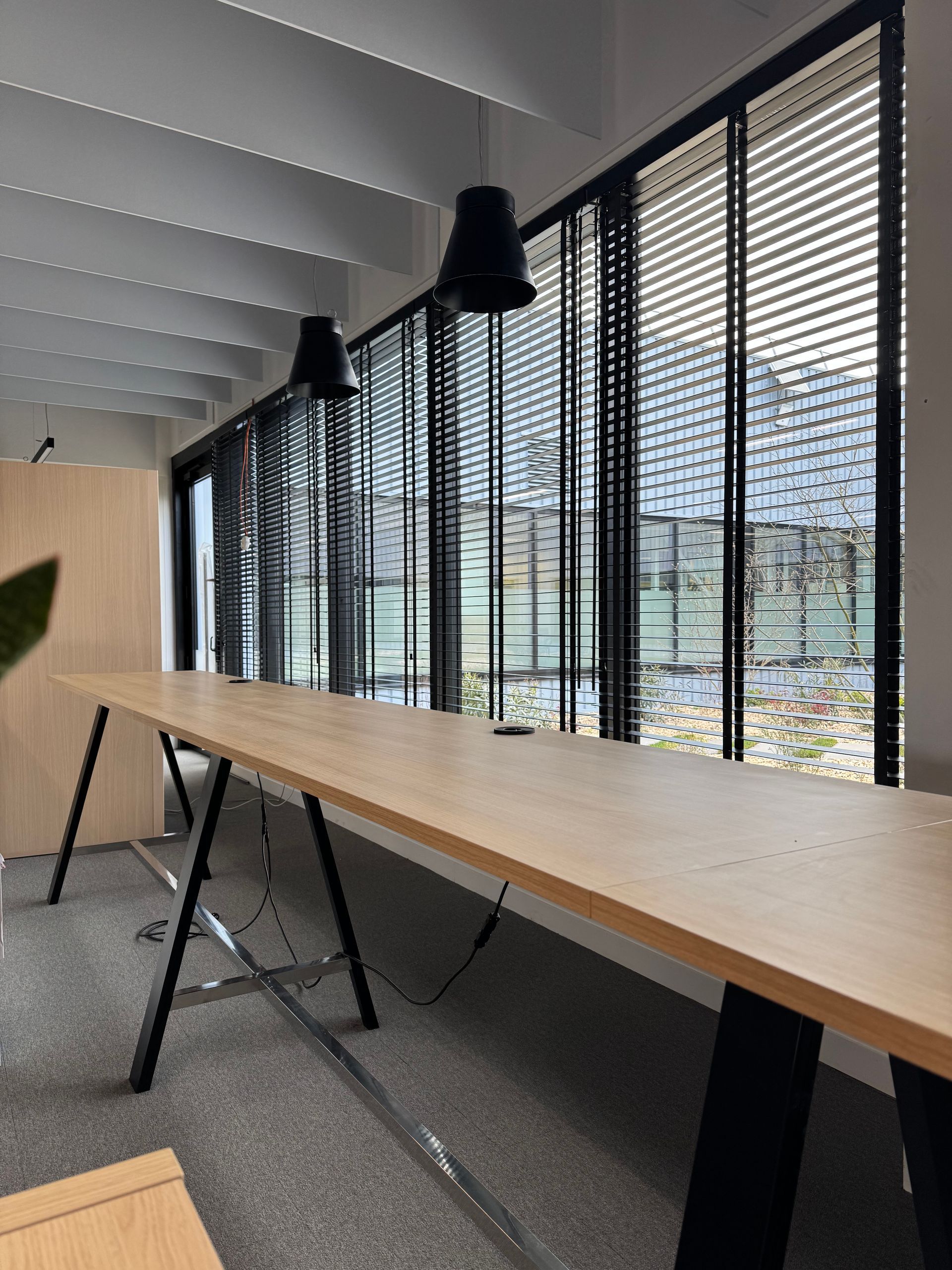 Une longue table en bois dans une pièce avec une grande fenêtre, deux lampes suspendues noires et un plafond clair.