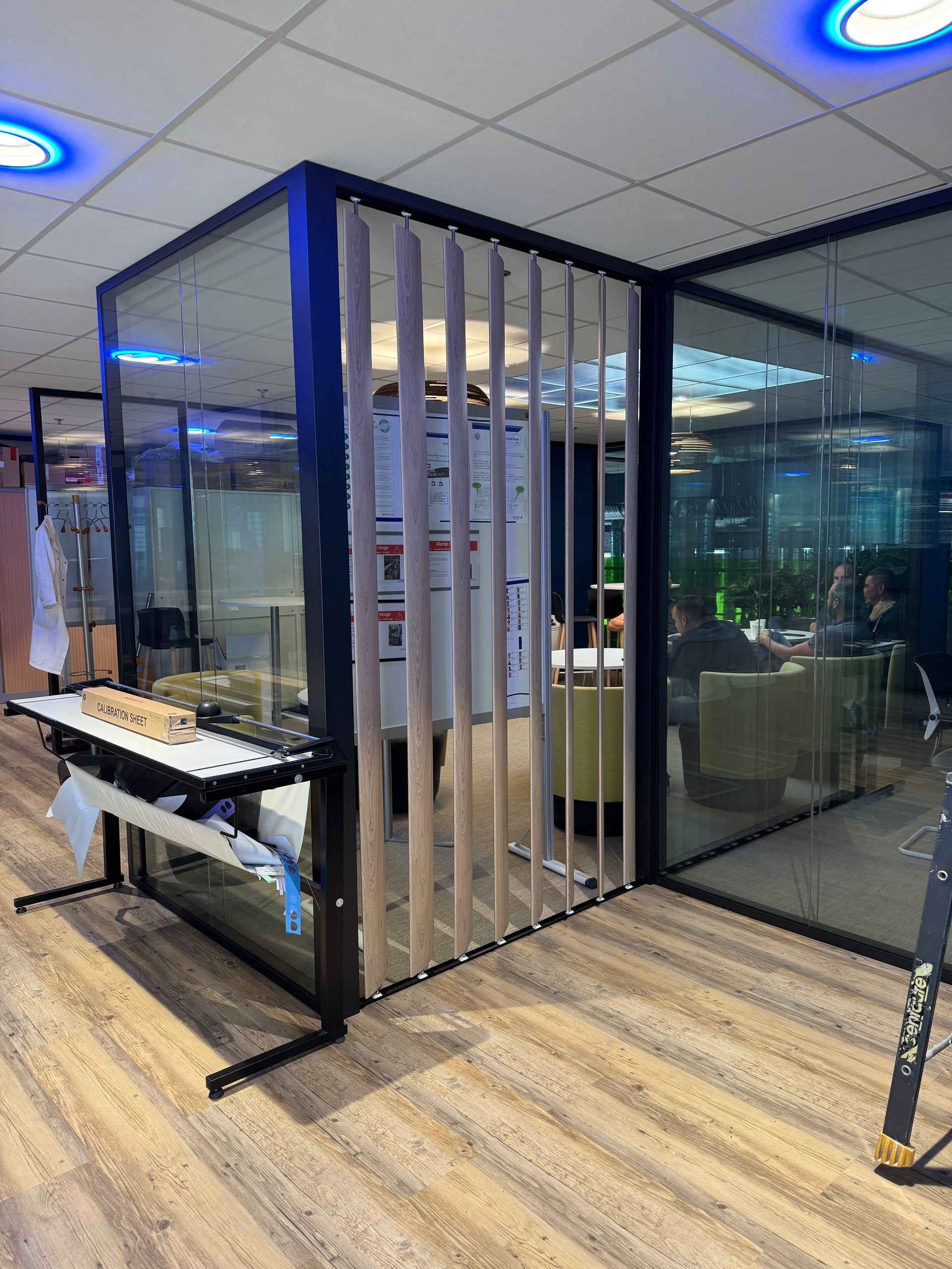 Espace de bureaux avec cloisons vitrées, parquet et stores verticaux. Des personnes sont assises à l'intérieur, des plans sont posés sur une table.