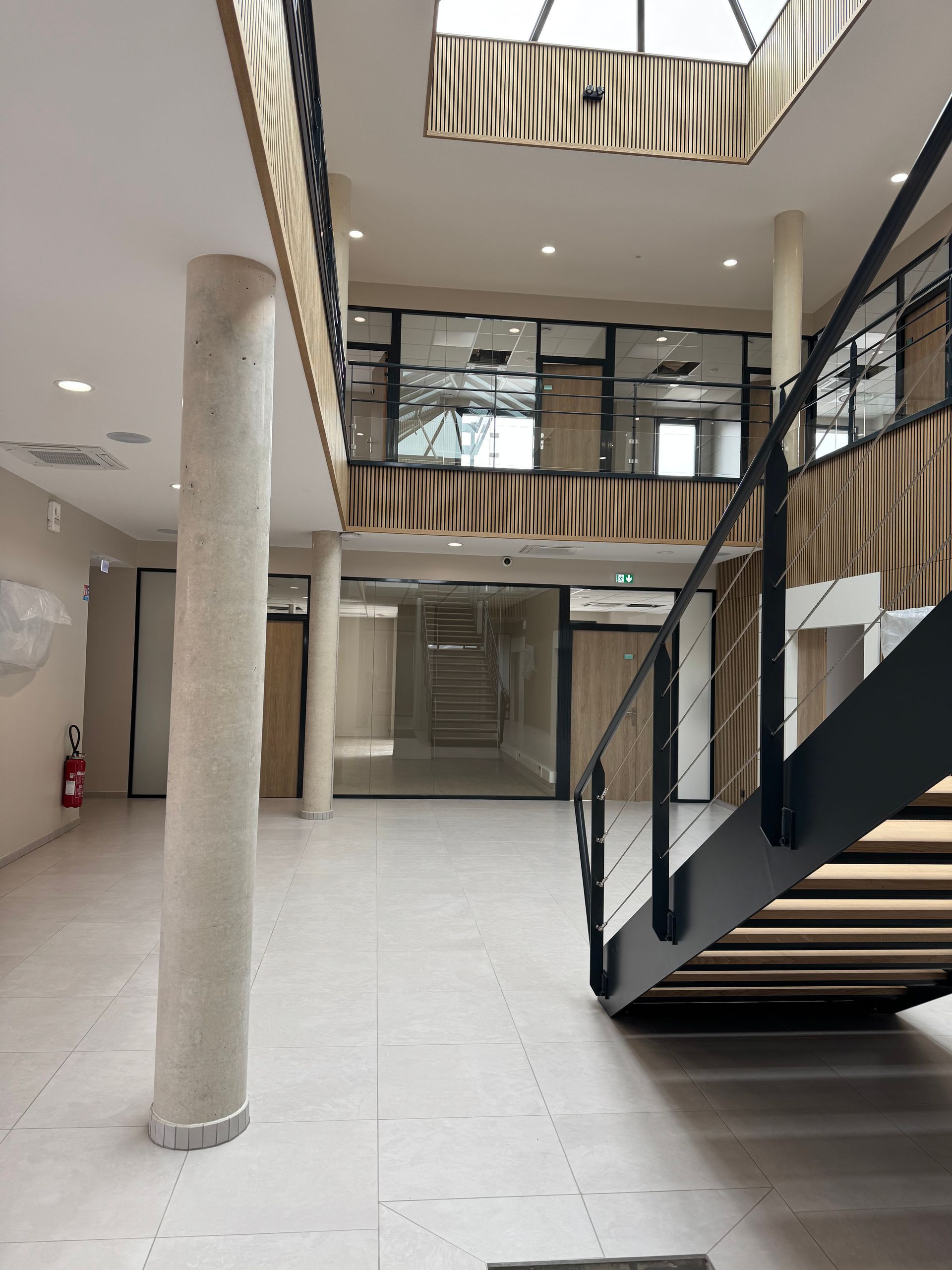 Vue intérieure d'un bâtiment moderne avec escalier, colonnes et pièces aux parois de verre, baignées de lumière naturelle.