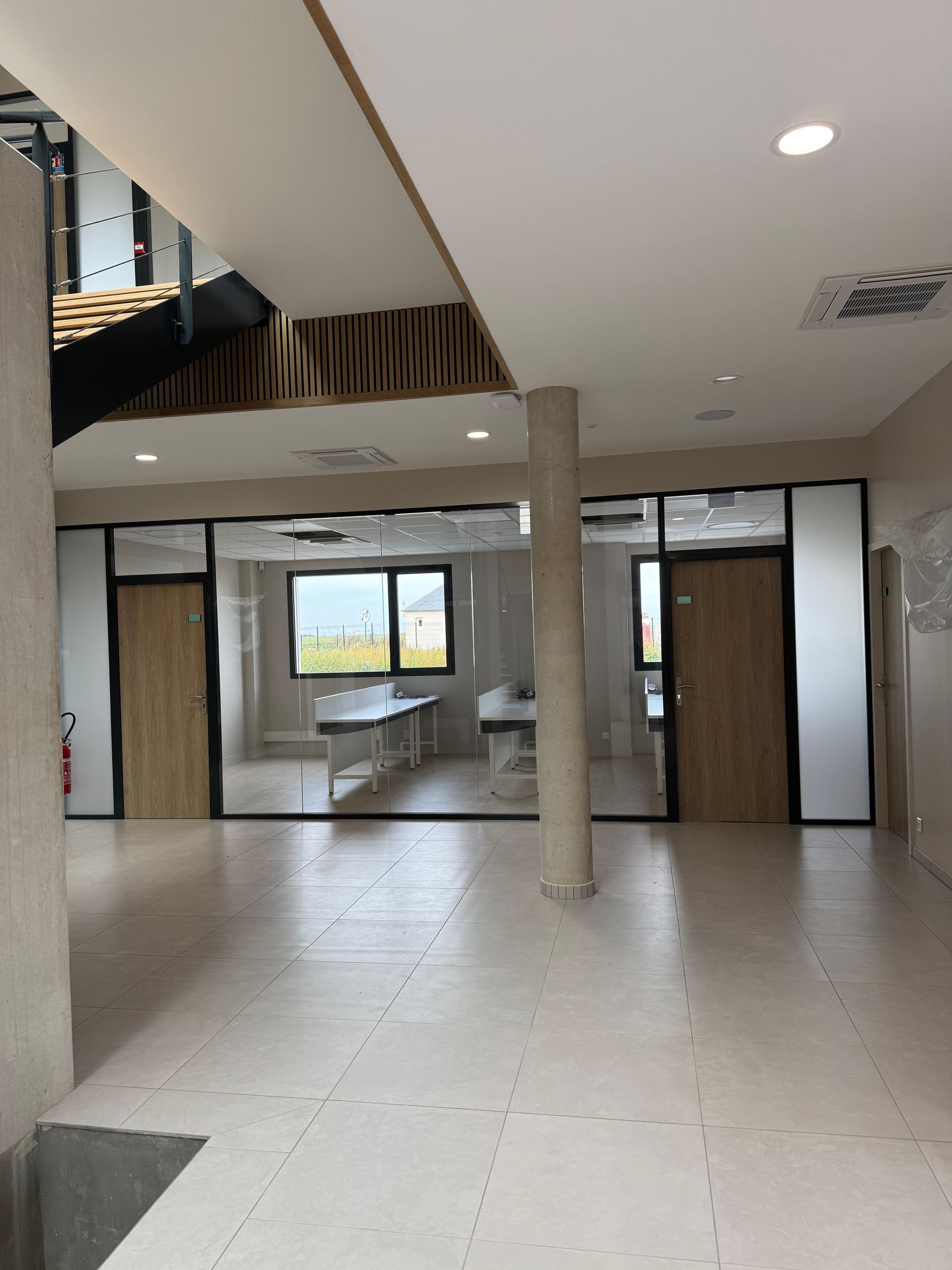 Couloir avec pièces vitrées, portes en bois, pilier en béton et escalier. Couleurs claires et neutres.